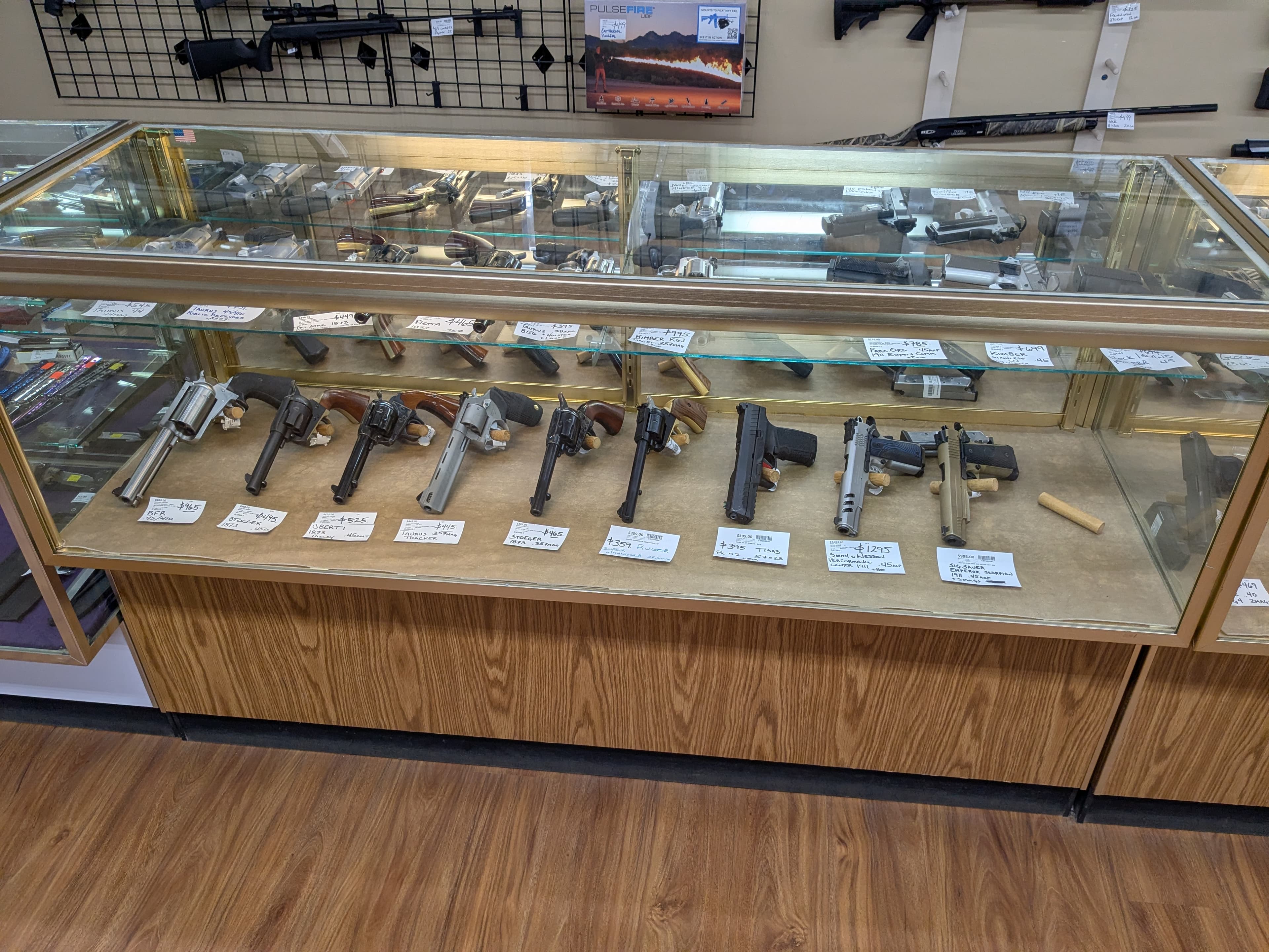 Glass display case with revolvers and semi-automatic handguns — rifles and shotguns on wall above