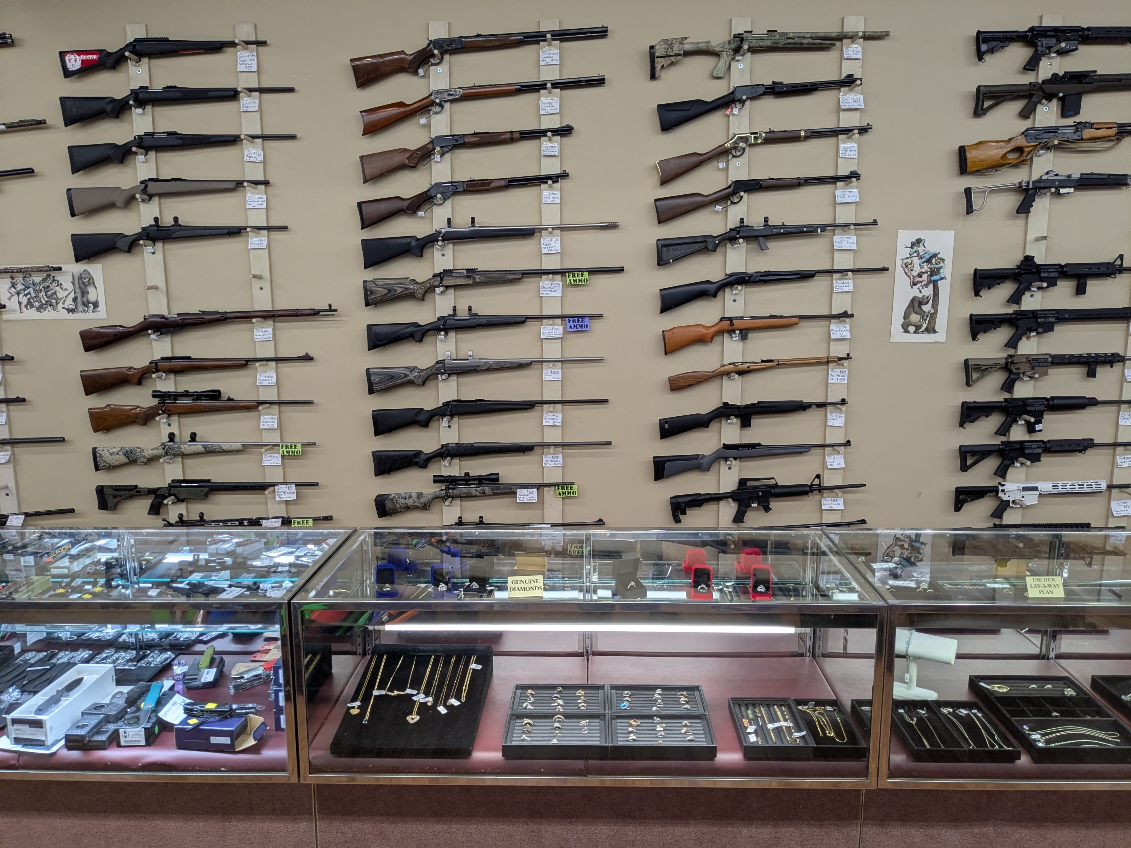 Wall of rifles and shotguns including bolt-action, AR-style, and scoped hunting rifles — jewelry and knife cases below