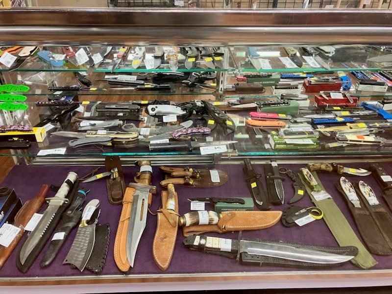 Glass display case with hunting knives, Bowie knives with leather sheaths, and folding knives on purple velvet